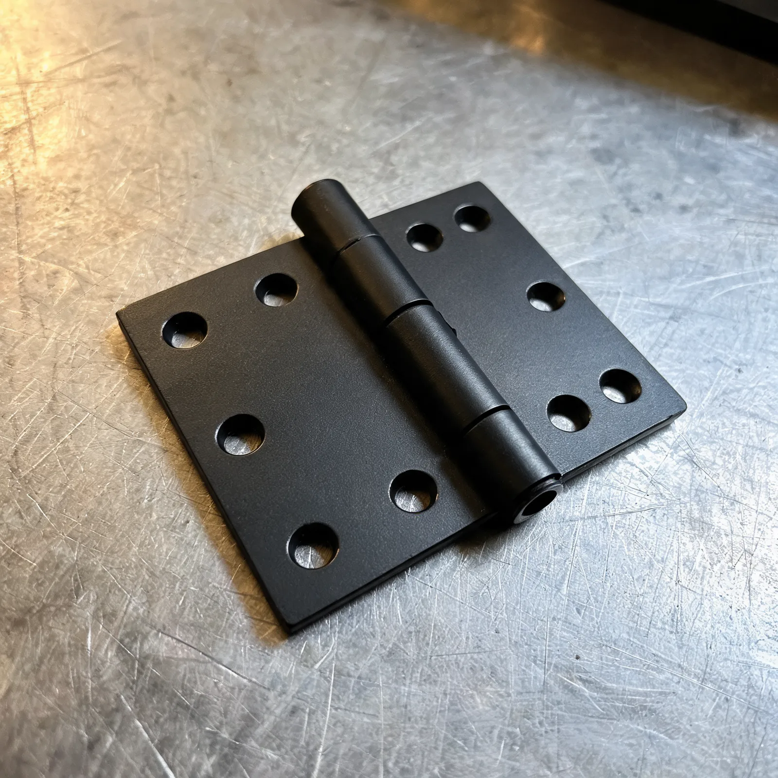 A matte black commercial hinge with squared leaves and visible screw holes on a steel bench.